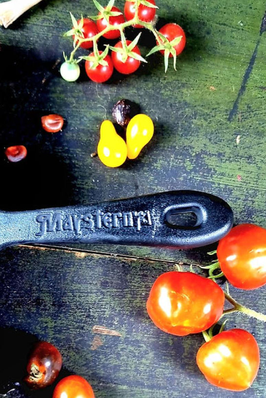 Tomatoes and a black spoon with 'Maysternya' branding on a dark surface