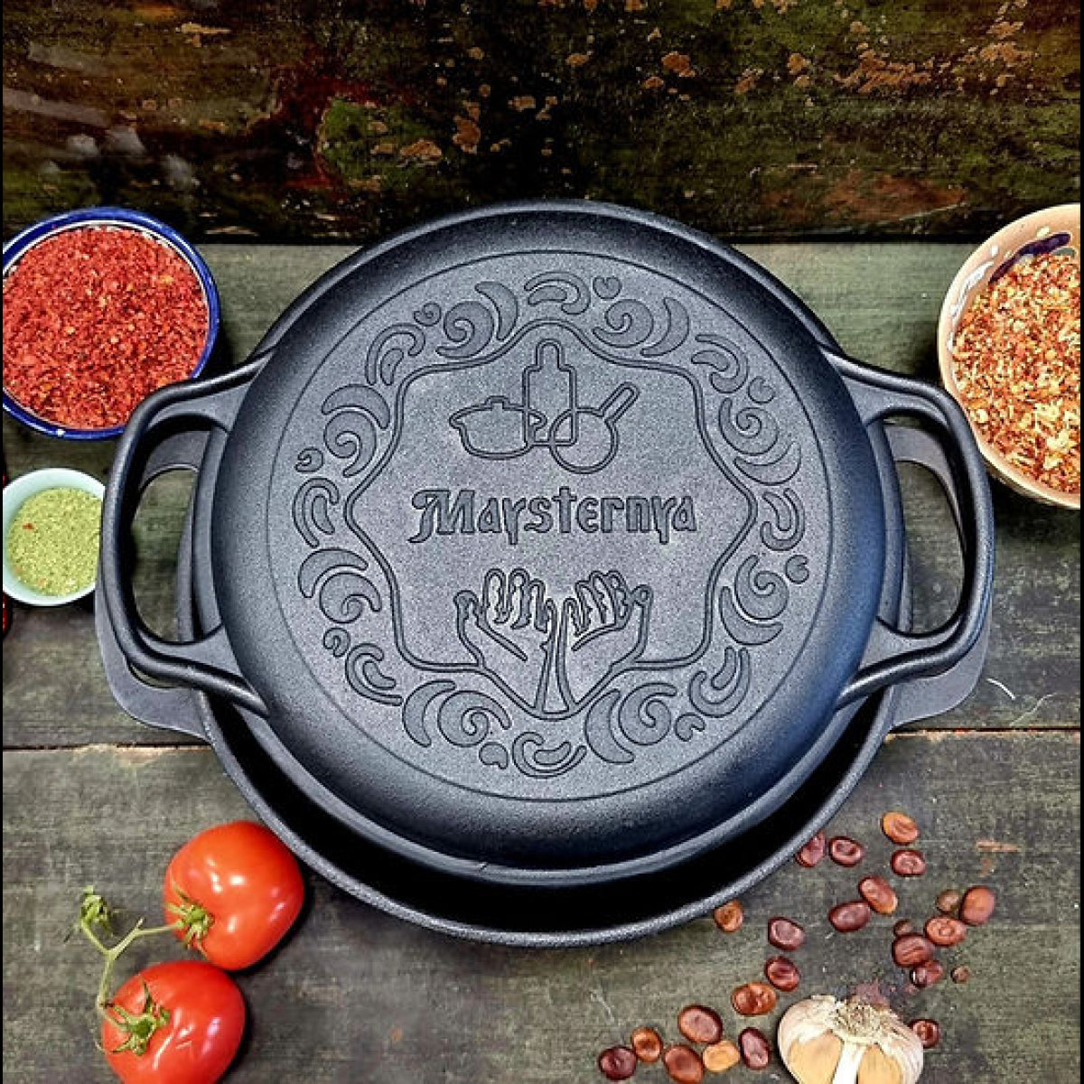 Black cast iron skillet with 'Maysternya' branding on a wooden surface with ingredients.