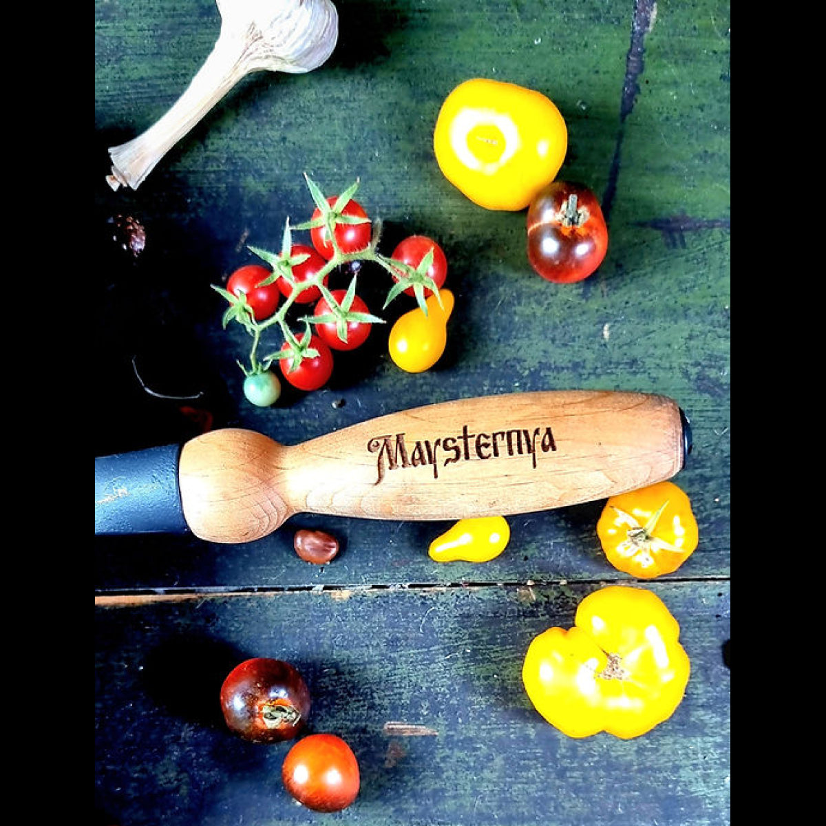 Wooden mace with 'Maysternya' branding surrounded by tomatoes and garlic on a dark surface