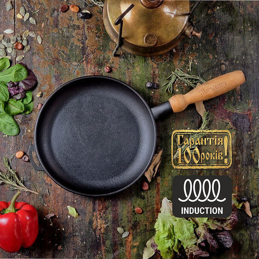 Black cast iron pancake pan with wooden handle on a rustic wooden surface with vegetables and a brand logo.