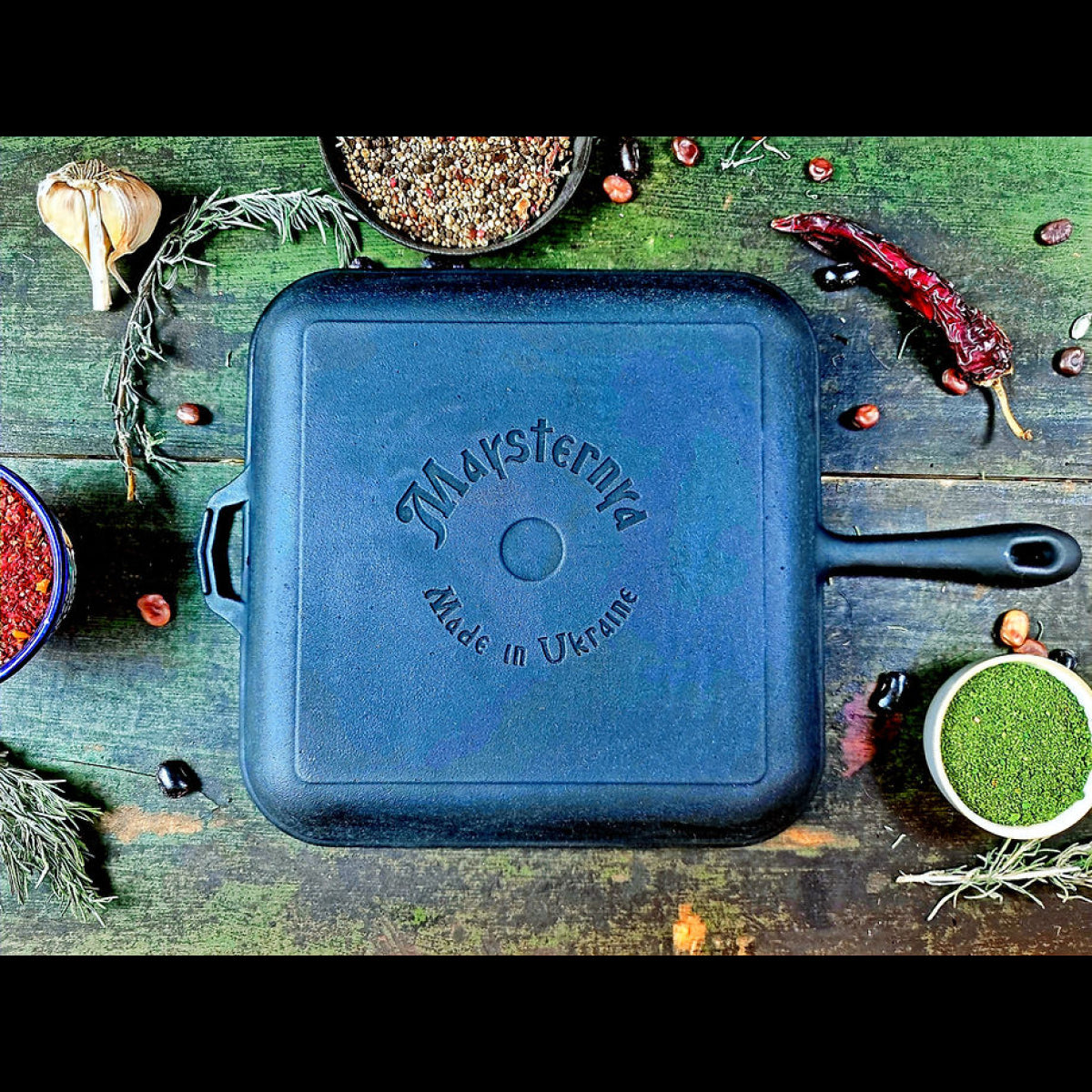 Black cast iron skillet with 'Maysternya' branding on a wooden surface with spices and herbs.