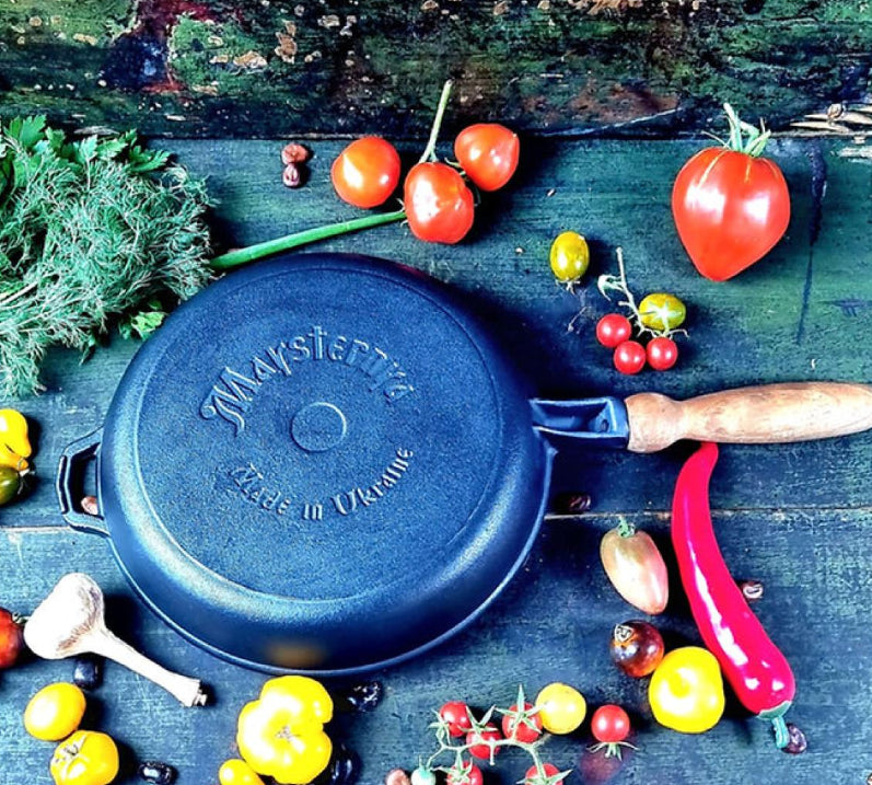 Black cast iron pan with 'Maysternya' branding on a wooden surface with vegetables.