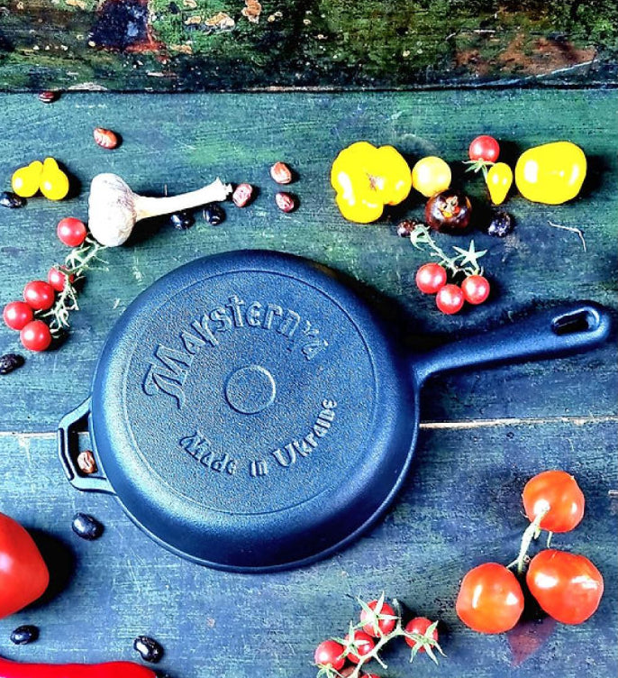 Blue cast iron skillet with 'Maysternya' branding on a rustic wooden surface with vegetables.