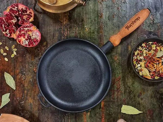 Black cast iron frying pan on a rustic wooden surface with pomegranate, spices, and a wooden handle.