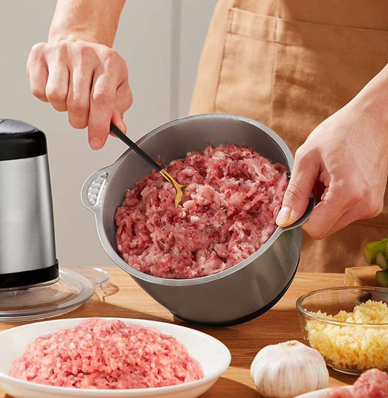Person handling ground meat in a bowl with a grinder and ingredients on a wooden surface.