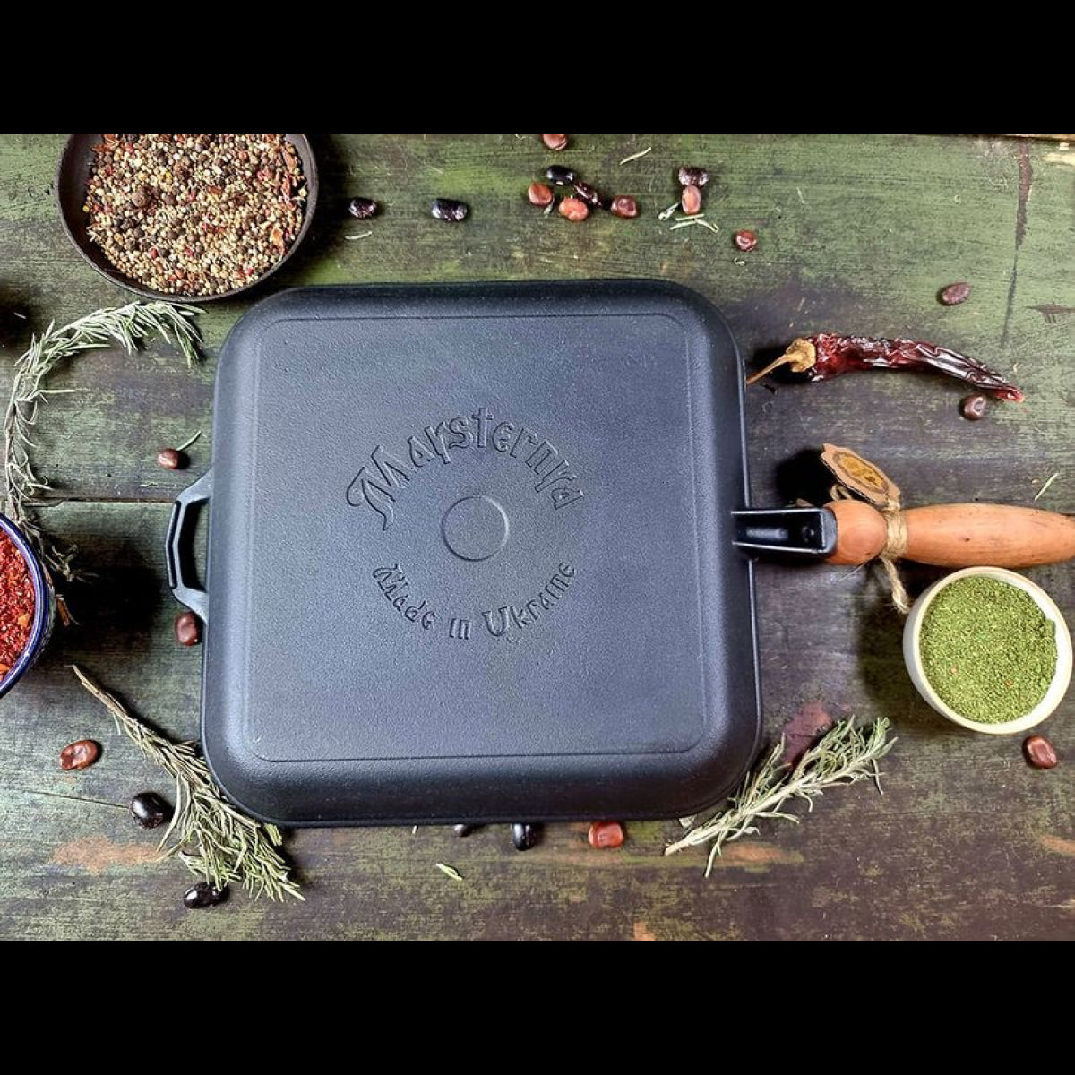 Black сast iron frying pan with 'Maysternya' branding on a rustic wooden surface with ingredients.
